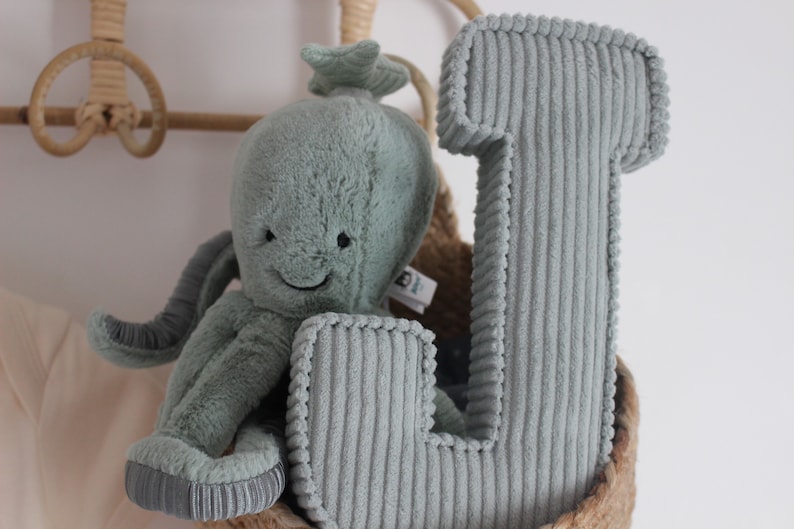 May include: A plush, light teal octopus toy with a smiling face and a corduroy letter "L" pillow are nestled in a woven basket. The octopus has eight tentacles and a star-shaped head. The letter is a soft, corduroy fabric.