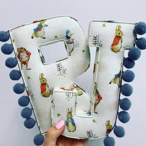 May include: A large, white fabric letter R with a Peter Rabbit print. The letter is decorated with blue pom-poms around the edges.