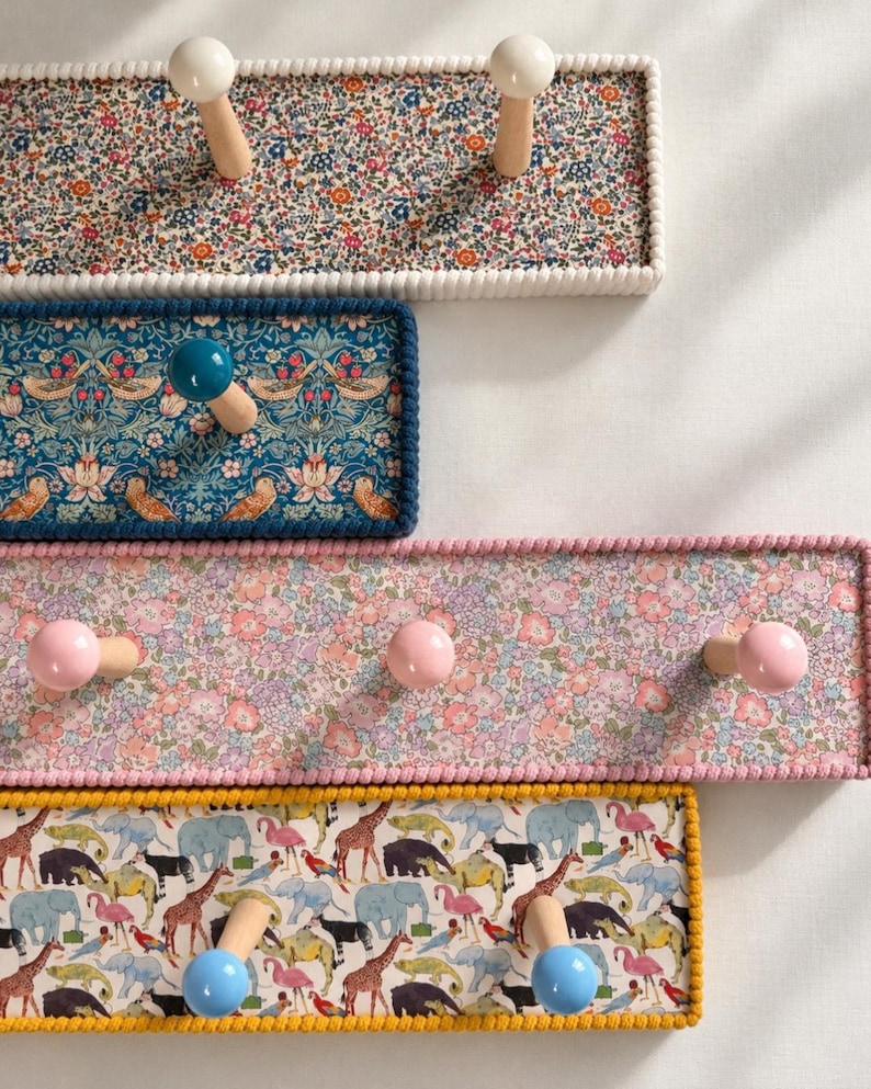 Puede incluir: Cuatro percheros de pared rectangulares con paneles cubiertos de tela. El estante superior tiene un estampado floral y pomos blancos. El segundo tiene un patr&oacute;n de p&aacute;jaros azules y un pomo azul. El tercero tiene un estampado floral rosa y pomos rosas. El inferior tiene un estampado de animales y pomos azules.