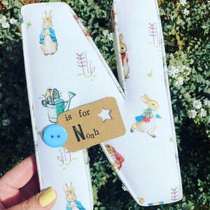 May include: A white fabric letter N with a Peter Rabbit design. The letter has a blue button and a brown tag that says "is for Noah".