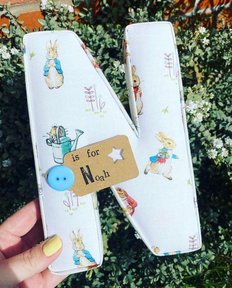 May include: A white fabric letter N with a Peter Rabbit design. The letter has a blue button and a brown tag that says "is for Noah".