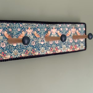 Puede incluir: Un perchero de pared rectangular con un panel de tela en color verde azulado oscuro con un patr&oacute;n repetitivo de p&aacute;jaros, flores y follaje. Incluye tres ganchos de madera con extremos redondeados en azul oscuro y un borde negro.