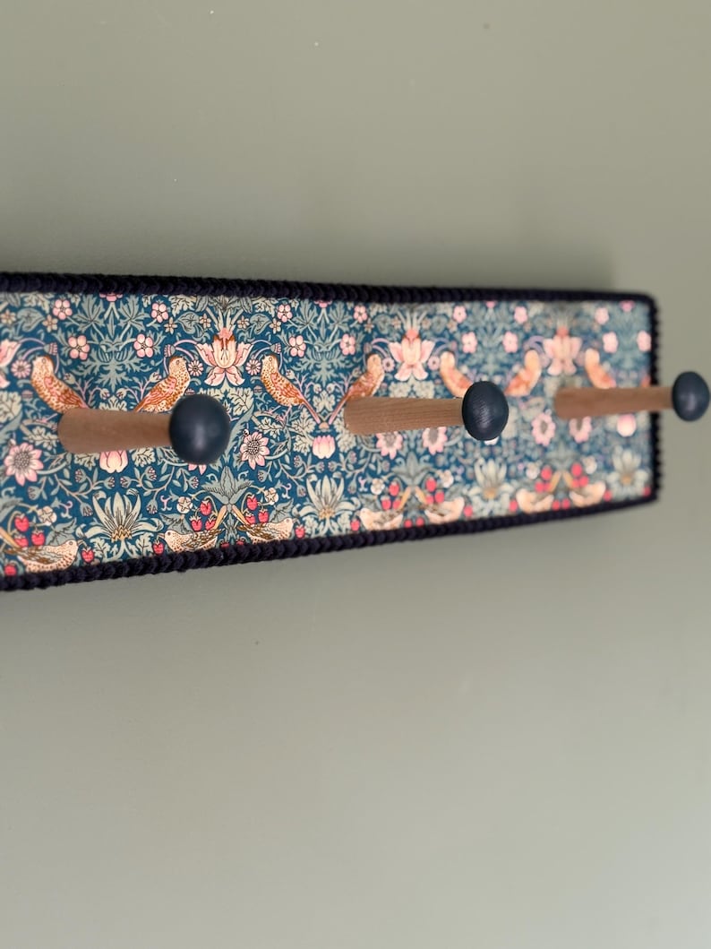 Puede incluir: Un perchero de pared rectangular con un panel de tela en color verde azulado oscuro con un patr&oacute;n repetitivo de p&aacute;jaros, flores y follaje. Incluye tres ganchos de madera con extremos redondeados en azul oscuro y un borde negro.