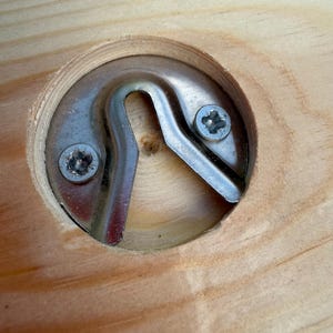 Puede incluir: Una placa de metal plateado para colgar, empotrada en un corte circular en una superficie de madera clara. El herraje est&aacute; asegurado con dos peque&ntilde;os tornillos de estrella y presenta una abertura central en forma de U.