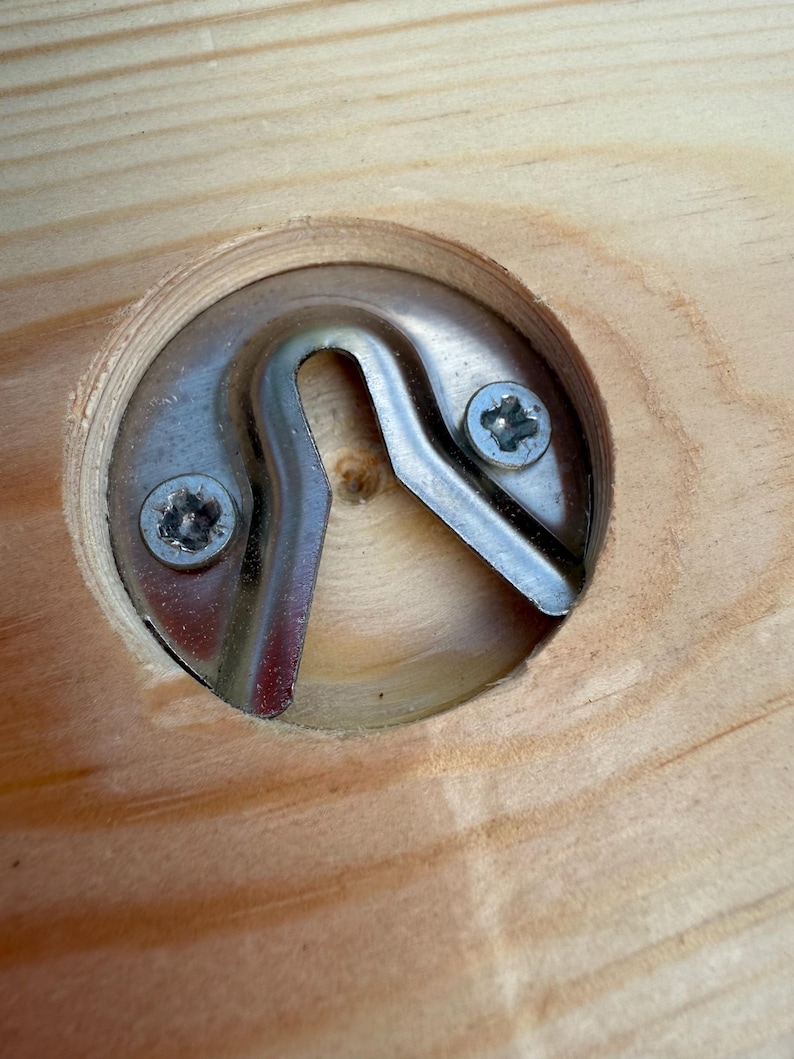 Puede incluir: Una placa de metal plateado para colgar, empotrada en un corte circular en una superficie de madera clara. El herraje est&aacute; asegurado con dos peque&ntilde;os tornillos de estrella y presenta una abertura central en forma de U.