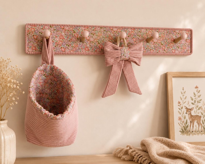 Puede incluir: Un perchero de pared de madera con incrustaciones de tela floral y cinco ganchos de color rosa claro. Una bolsa de almacenamiento colgante de tela con un interior floral a juego y un lazo de tela acanalada rosa cuelgan de los ganchos.