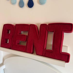 May include: A red corduroy BEN name sign displayed on a white shelf. Above the sign, a garland of blue felt raindrops hangs against a white wall. The letters are thick and plush, creating a 3D effect.