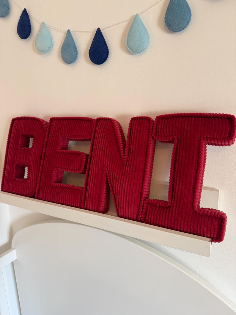 May include: A red corduroy BEN name sign displayed on a white shelf. Above the sign, a garland of blue felt raindrops hangs against a white wall. The letters are thick and plush, creating a 3D effect.