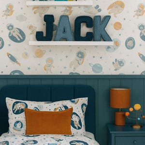 May include: A child's bedroom with a space theme. The bed has a white comforter with a space-themed pattern and an orange pillow. The wall features a wallpaper with astronauts, planets, and rockets. The name "JACK" is displayed on the wall.