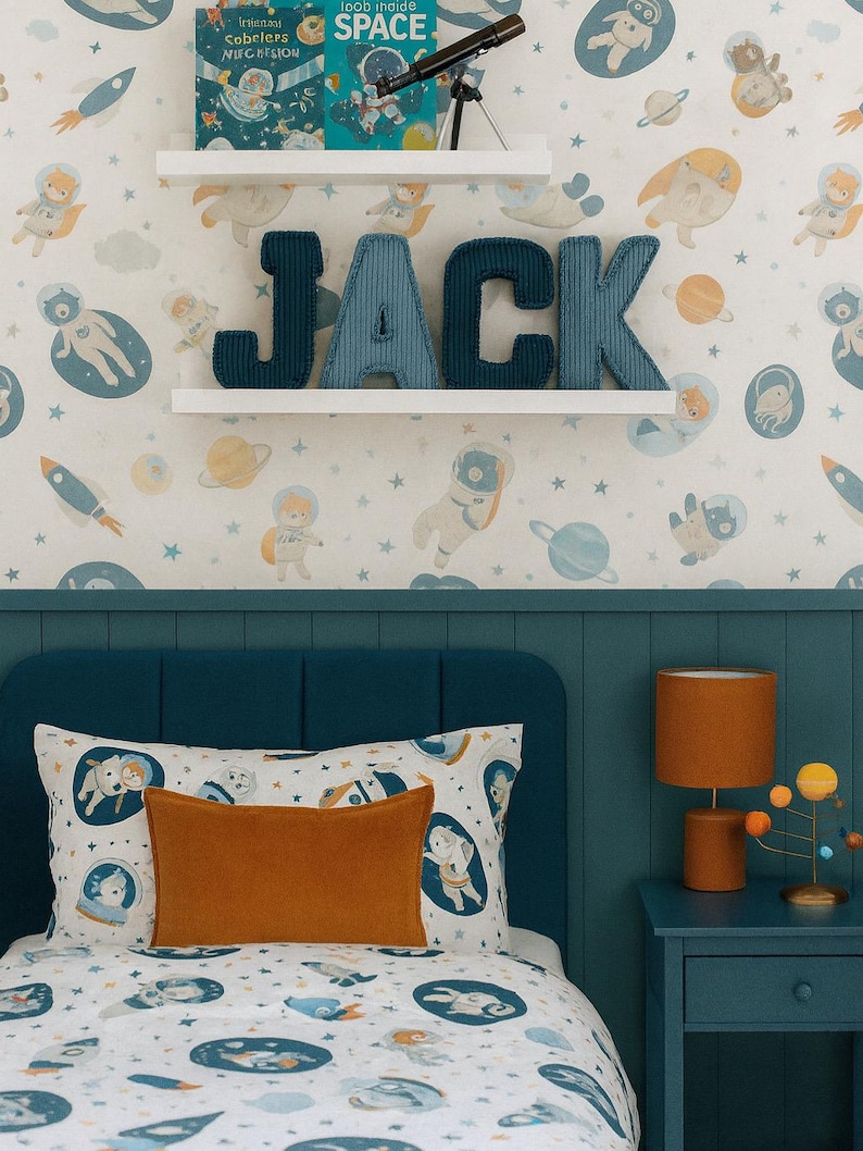 May include: A child's bedroom with a space theme. The bed has a white comforter with a space-themed pattern and an orange pillow. The wall features a wallpaper with astronauts, planets, and rockets. The name "JACK" is displayed on the wall.