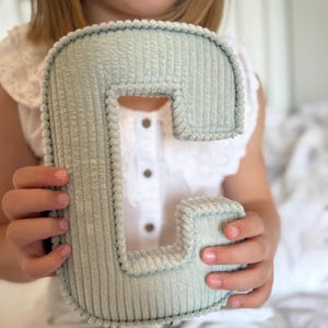 May include: A soft, corduroy letter "C" in a muted green color. The plush letter is held by a person, showcasing its textured surface and rounded edges. The letter is a decorative item, possibly for a nursery or children's room.