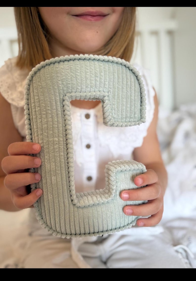 May include: A soft, corduroy letter "C" in a muted green color. The plush letter is held by a person, showcasing its textured surface and rounded edges. The letter is a decorative item, possibly for a nursery or children's room.