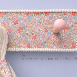 Puede incluir: Un perchero de pared rectangular con un estampado floral en tonos rosa, azul y amarillo, bordeado con un adorno de pompones blancos. Incluye dos ganchos redondos de madera rosa y el texto: Psst. have you ever seen a peg rack this beautiful?