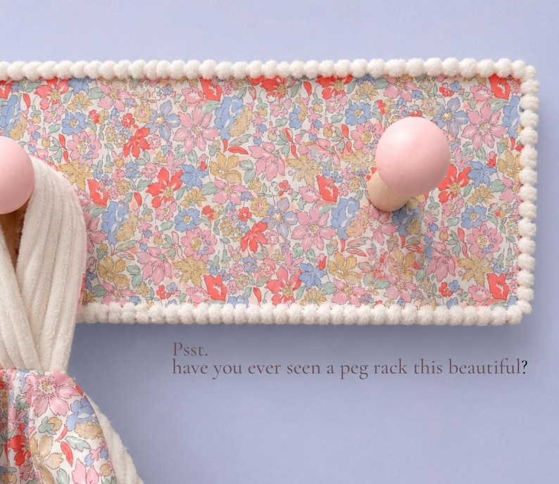 Puede incluir: Un perchero de pared rectangular con un estampado floral en tonos rosa, azul y amarillo, bordeado con un adorno de pompones blancos. Incluye dos ganchos redondos de madera rosa y el texto: Psst. have you ever seen a peg rack this beautiful?