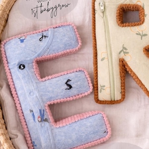 May include: Close-up of two fabric letters, "E" and "R", made from baby clothes. The "E" is light blue with pink trim and embroidered details. The "R" is beige with brown trim. Text reads "handmade with their 1st babygrow" and "A keepsake made from the babygrow you just can't bare to part with."