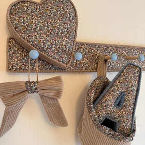 Puede incluir: Un perchero de pared con ganchos azul claro, con un panel de tela floral y un coj&iacute;n a juego en forma de coraz&oacute;n. Un lazo de pana beige cuelga de un gancho, y una cesta de almacenamiento de pana beige con interior floral cuelga de otro.