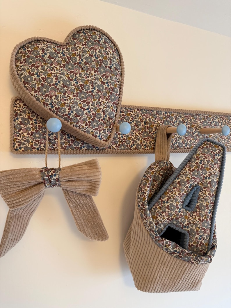 Puede incluir: Un perchero de pared con ganchos azul claro, con un panel de tela floral y un coj&iacute;n a juego en forma de coraz&oacute;n. Un lazo de pana beige cuelga de un gancho, y una cesta de almacenamiento de pana beige con interior floral cuelga de otro.