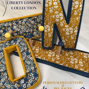 Puede incluir: Una letra O y una letra N decorativas, y un perchero, todos cubiertos de tela floral con ribetes de pana en color amarillo mostaza y azul marino. El texto dice THE LIBERTY LONDON COLLECTION y PERSONALISED LETTERS & PEG RACKS MATCHY MATCHY.