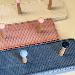 Può includere: Primo piano di tre attaccapanni a parete in velluto a coste bianchi, rosa e blu. Ogni attaccapanni ha pioli in legno con cime colorate in rosa, giallo, blu e nero. Gli attaccapanni sono rettangolari con una superficie strutturata.
