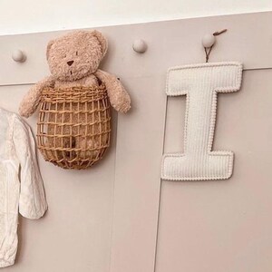 May include: A beige teddy bear sits in a woven basket hanging on a wall. A white, corduroy letter "I" is suspended from a small wooden peg. The wall is painted a light pink color, and there are several small, round pegs.