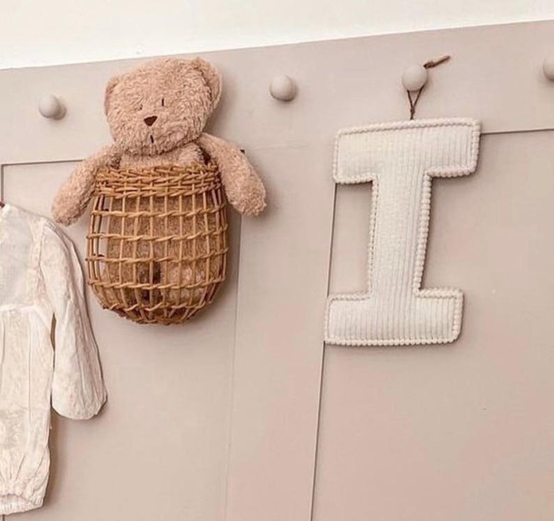 May include: A beige teddy bear sits in a woven basket hanging on a wall. A white, corduroy letter "I" is suspended from a small wooden peg. The wall is painted a light pink color, and there are several small, round pegs.