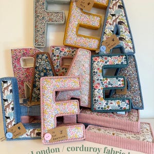 May include: Fabric letters in floral and animal prints, with corduroy borders. Each letter is a different colour. Some letters have tags with names. The letters are stacked. The text at the bottom reads "Liberty London / corduroy fabric letters."