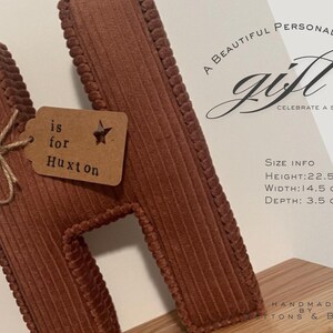 May include: A brown corduroy letter "H" with a tag that reads "is for Huxton". The letter is 8.9 inches tall, 5.7 inches wide, and 1.4 inches deep. The image also includes the text "A BEAUTIFUL PERSONALISED BABY GIFT CELEBRATE A SPECIAL NAME".