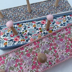 Puede incluir: Un perchero de pared con tres paneles horizontales cubiertos con telas de motivos florales. Los paneles est&aacute;n bordeados con trenzas y cuentan con perchas de madera, incluyendo dos pintadas de rosa claro, para colgar objetos.