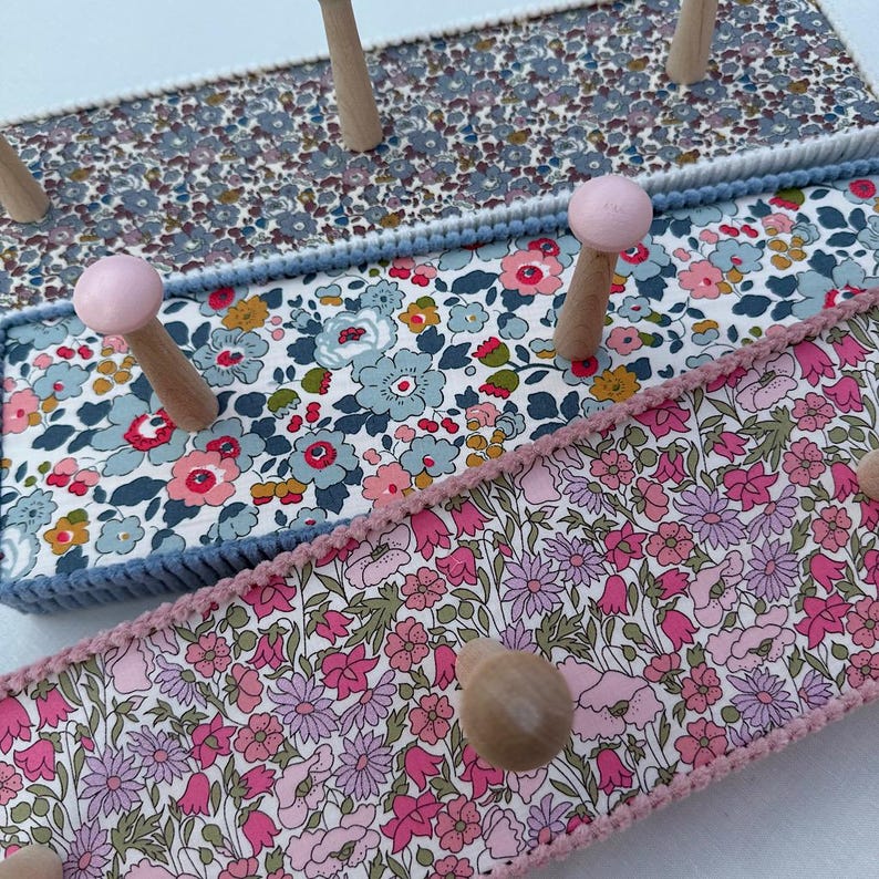Puede incluir: Un perchero de pared con tres paneles horizontales cubiertos con telas de motivos florales. Los paneles est&aacute;n bordeados con trenzas y cuentan con perchas de madera, incluyendo dos pintadas de rosa claro, para colgar objetos.
