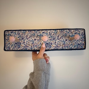 Puede incluir: Un perchero de pared rectangular con un patr&oacute;n de tela floral en tonos azules, morados y blancos. El perchero incluye tres ganchos de madera de color rosa claro y est&aacute; acabado con un borde decorativo azul oscuro en los bordes exteriores.