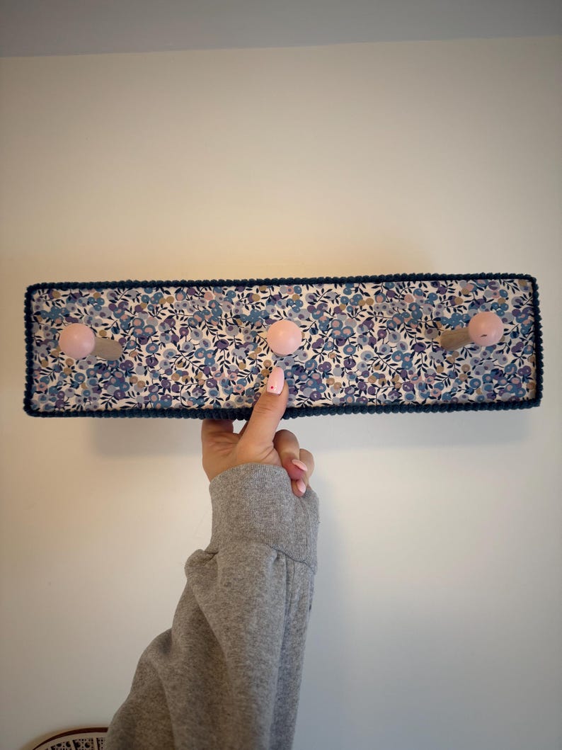 Puede incluir: Un perchero de pared rectangular con un patr&oacute;n de tela floral en tonos azules, morados y blancos. El perchero incluye tres ganchos de madera de color rosa claro y est&aacute; acabado con un borde decorativo azul oscuro en los bordes exteriores.