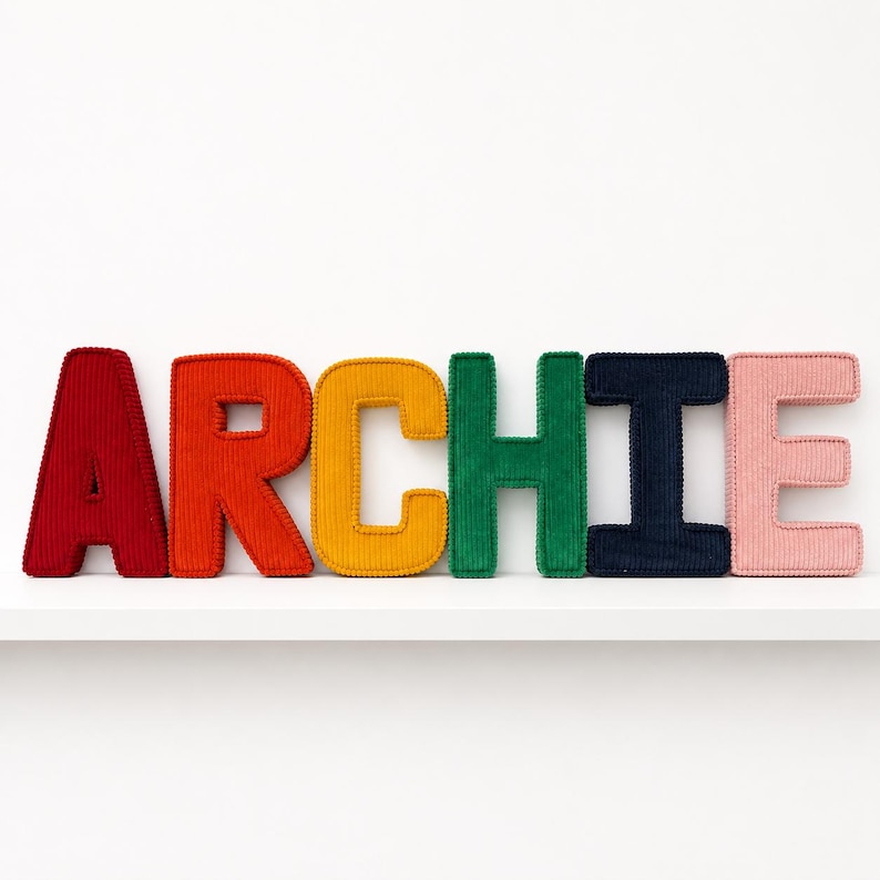 May include: A colorful display of the name "ARCHIE" spelled out in plush, corduroy letters. Each letter is a different color: red, orange, yellow, green, navy, and pink. The letters are arranged on a white shelf against a white background.
