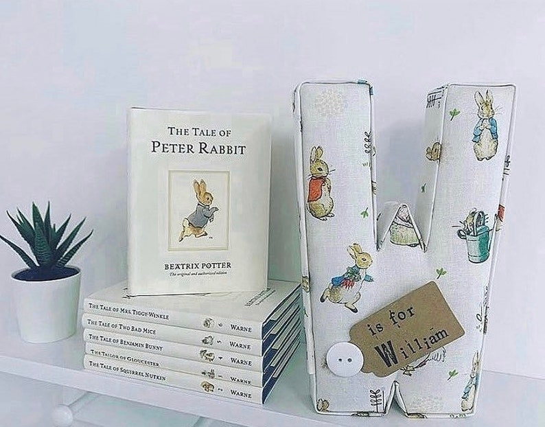 May include: A white fabric letter W with a Peter Rabbit design. The letter is decorated with a brown tag that says "is for William". The letter is sitting on a shelf with a stack of Peter Rabbit books.