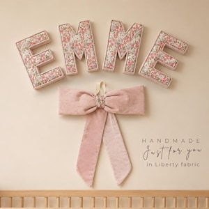 May include: Four padded letters spelling EMME, each covered in a floral fabric with pink, red, and green blossoms. Below the letters is a large, dusty pink corduroy bow with a floral center. Text reads: HANDMADE Just for you in Liberty fabric.