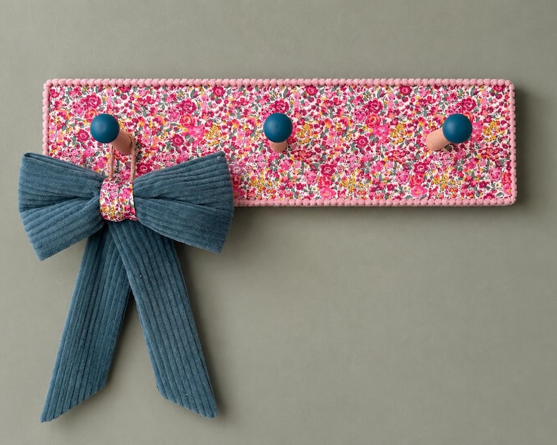 Puede incluir: Un perchero de pared rectangular con un estampado floral en tonos rosa y multicolor, bordeado por peque&ntilde;as cuentas rosas. Incluye tres ganchos de madera con extremos en azul cerceta, con un lazo de pana azul cerceta colgado de uno.