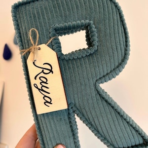 May include: A teal corduroy letter "R" with a wooden tag that says "Raya". The letter has a textured surface and a stitched border. The tag is tied with twine. Decorative item.