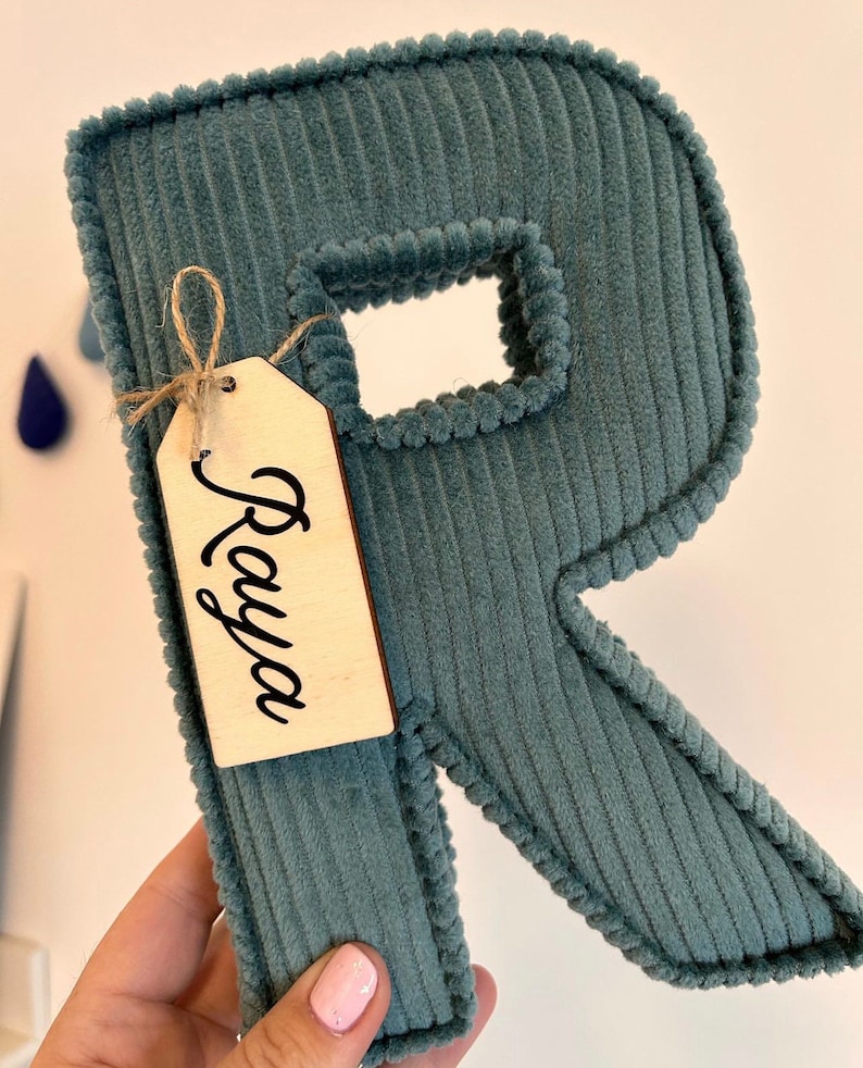 May include: A teal corduroy letter "R" with a wooden tag that says "Raya". The letter has a textured surface and a stitched border. The tag is tied with twine. Decorative item.
