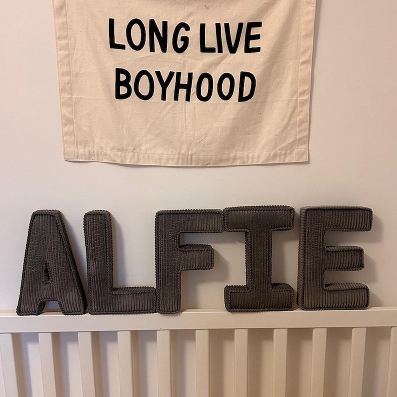 May include: A banner with the text "LONG LIVE BOYHOOD" hangs above the name "ALFIE" spelled out in gray corduroy letters. The letters are placed on a white crib rail. The banner and letters are against a white wall.