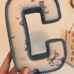 May include: A blue felt letter C with a white fabric background featuring a Peter Rabbit pattern. The letter is hand-stitched with a blue cord around the edges.