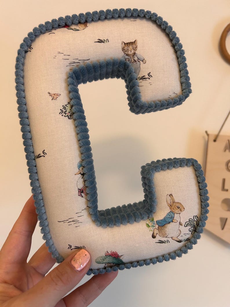 May include: A blue felt letter C with a white fabric background featuring a Peter Rabbit pattern. The letter is hand-stitched with a blue cord around the edges.