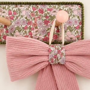 Puede incluir: Un lazo de tela acanalada rosa con un nudo central floral cuelga de un gancho de madera en un perchero de pared rectangular. El perchero cuenta con un panel de tela floral con flores rosas y verdes, acabado con un borde de pompones verde oscuro.