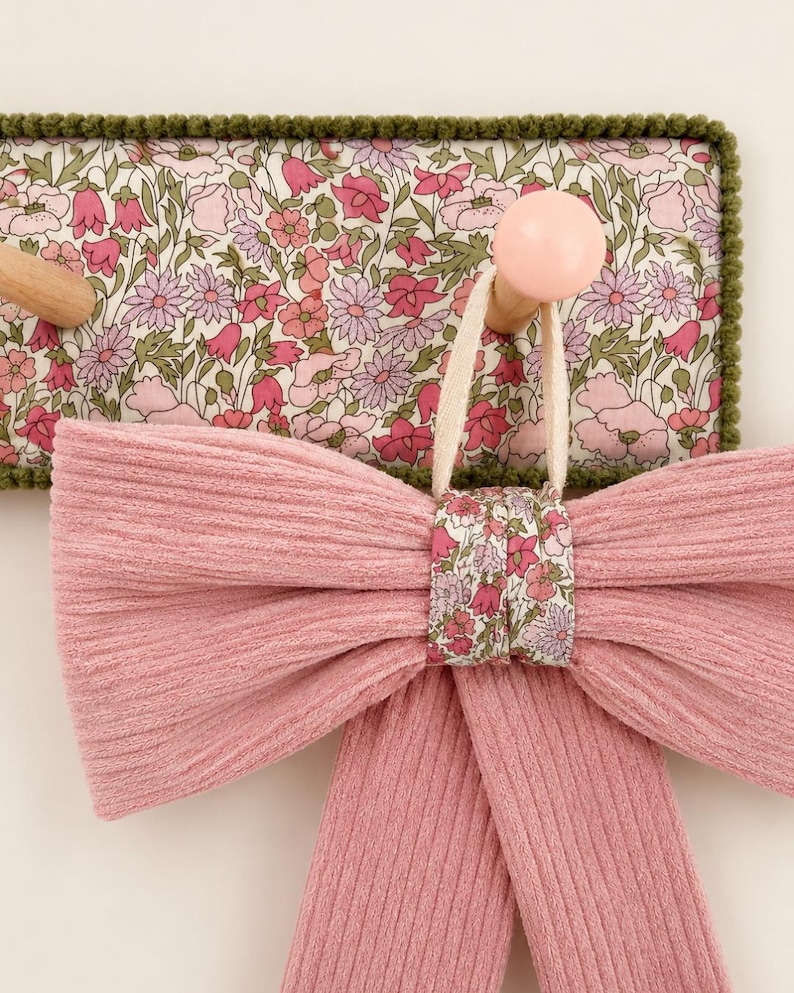 Puede incluir: Un lazo de tela acanalada rosa con un nudo central floral cuelga de un gancho de madera en un perchero de pared rectangular. El perchero cuenta con un panel de tela floral con flores rosas y verdes, acabado con un borde de pompones verde oscuro.
