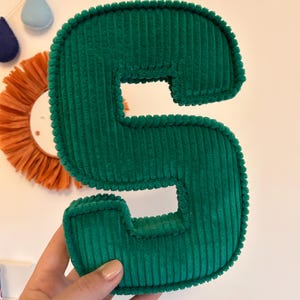 May include: A plush, emerald green corduroy letter "S" is held in a hand. The letter is thick and padded, with a textured surface. In the background are decorative wall hangings, including a sun-shaped piece and teardrop shapes.