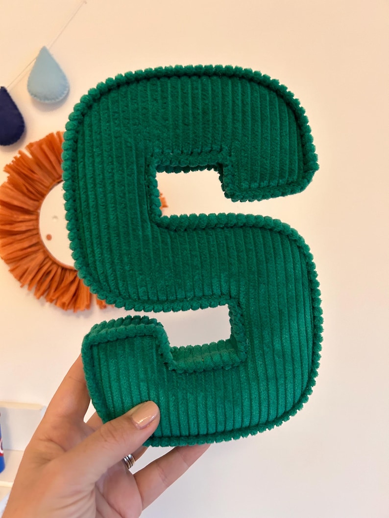 May include: A plush, emerald green corduroy letter "S" is held in a hand. The letter is thick and padded, with a textured surface. In the background are decorative wall hangings, including a sun-shaped piece and teardrop shapes.