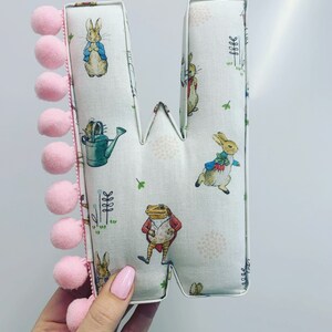 May include: A pink and white fabric letter W with a Peter Rabbit design. The letter is decorated with a pink pom-pom trim.