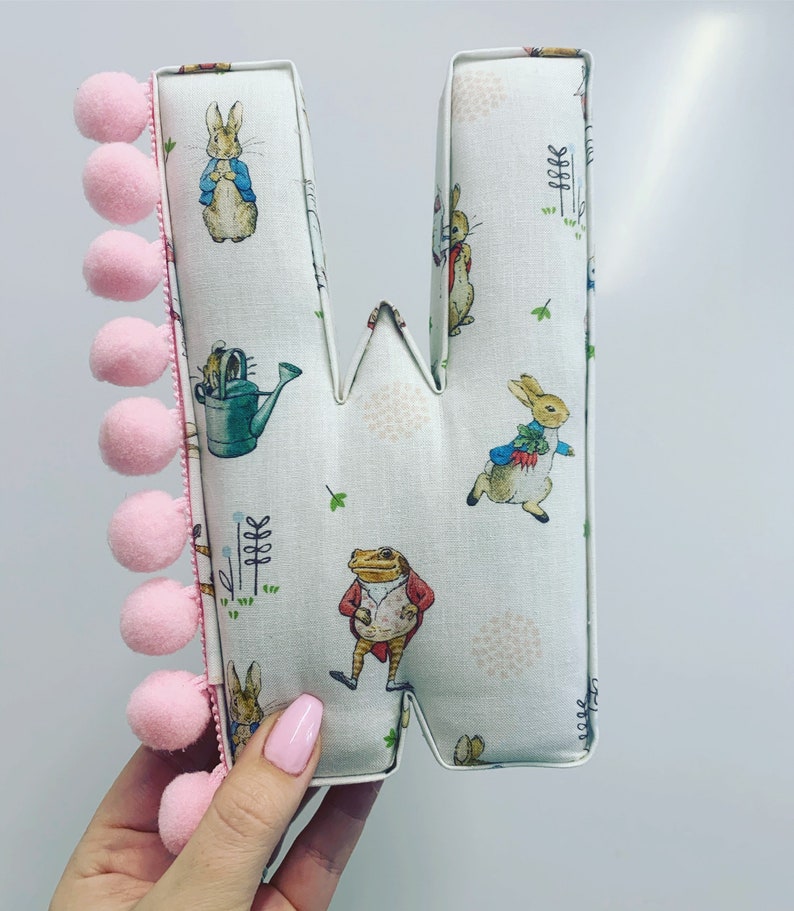 May include: A pink and white fabric letter W with a Peter Rabbit design. The letter is decorated with a pink pom-pom trim.