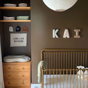 May include: A gold crib with a white sheet and a stuffed animal. The wall is painted a dark brown color and has a white paper lantern hanging from the ceiling. The wall also has the name "KAI" spelled out in felt letters. A wooden shelf with baskets and a sign that says "Long Live Boyhood" is on the left side of the image.