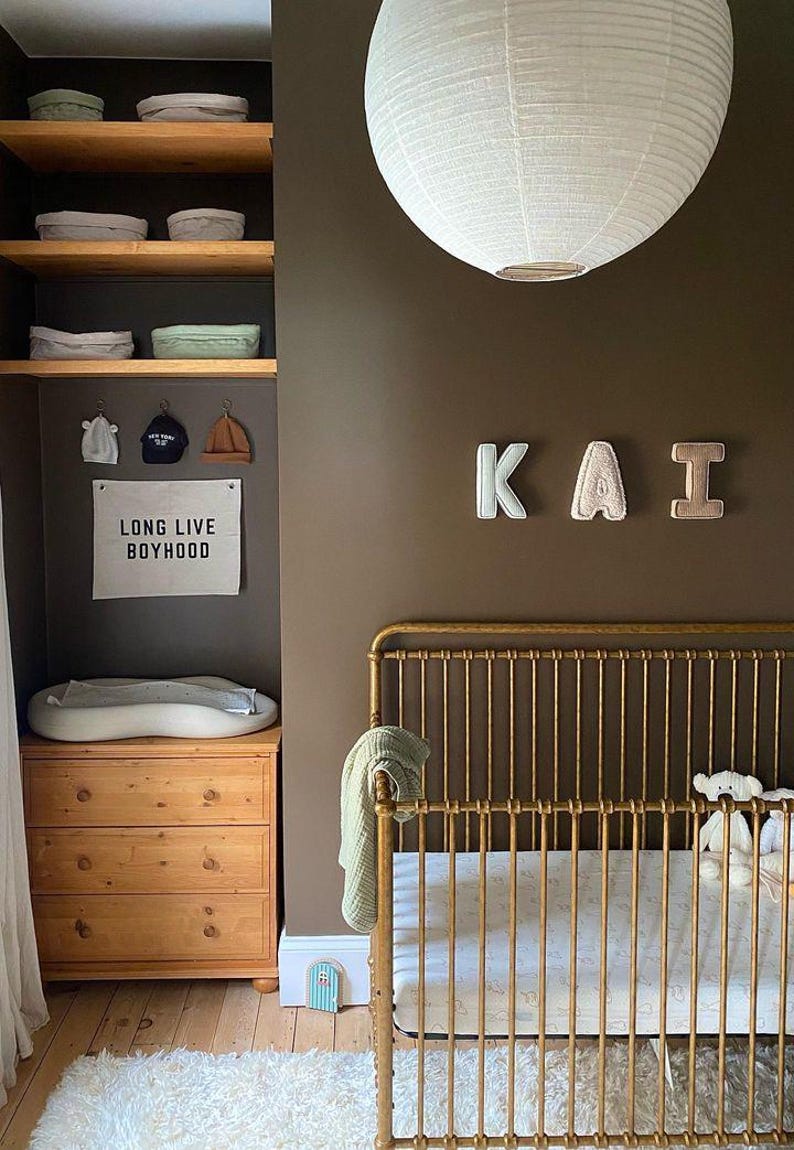 May include: A gold crib with a white sheet and a stuffed animal. The wall is painted a dark brown color and has a white paper lantern hanging from the ceiling. The wall also has the name "KAI" spelled out in felt letters. A wooden shelf with baskets and a sign that says "Long Live Boyhood" is on the left side of the image.