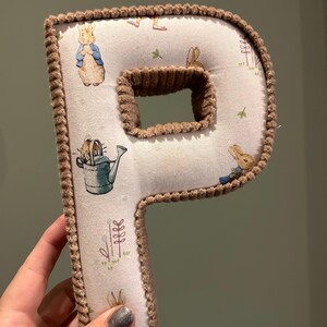 May include: A brown plush letter P with a white fabric background featuring a repeating pattern of Peter Rabbit characters. The letter is standing upright.