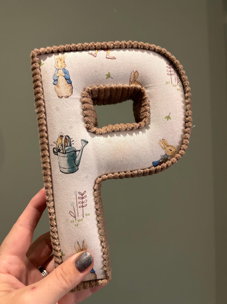 May include: A brown plush letter P with a white fabric background featuring a repeating pattern of Peter Rabbit characters. The letter is standing upright.